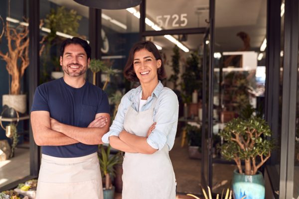 Portrait Of Smiling Male And Female Owners Of Florists Standing In Doorway Surrounded By Plants